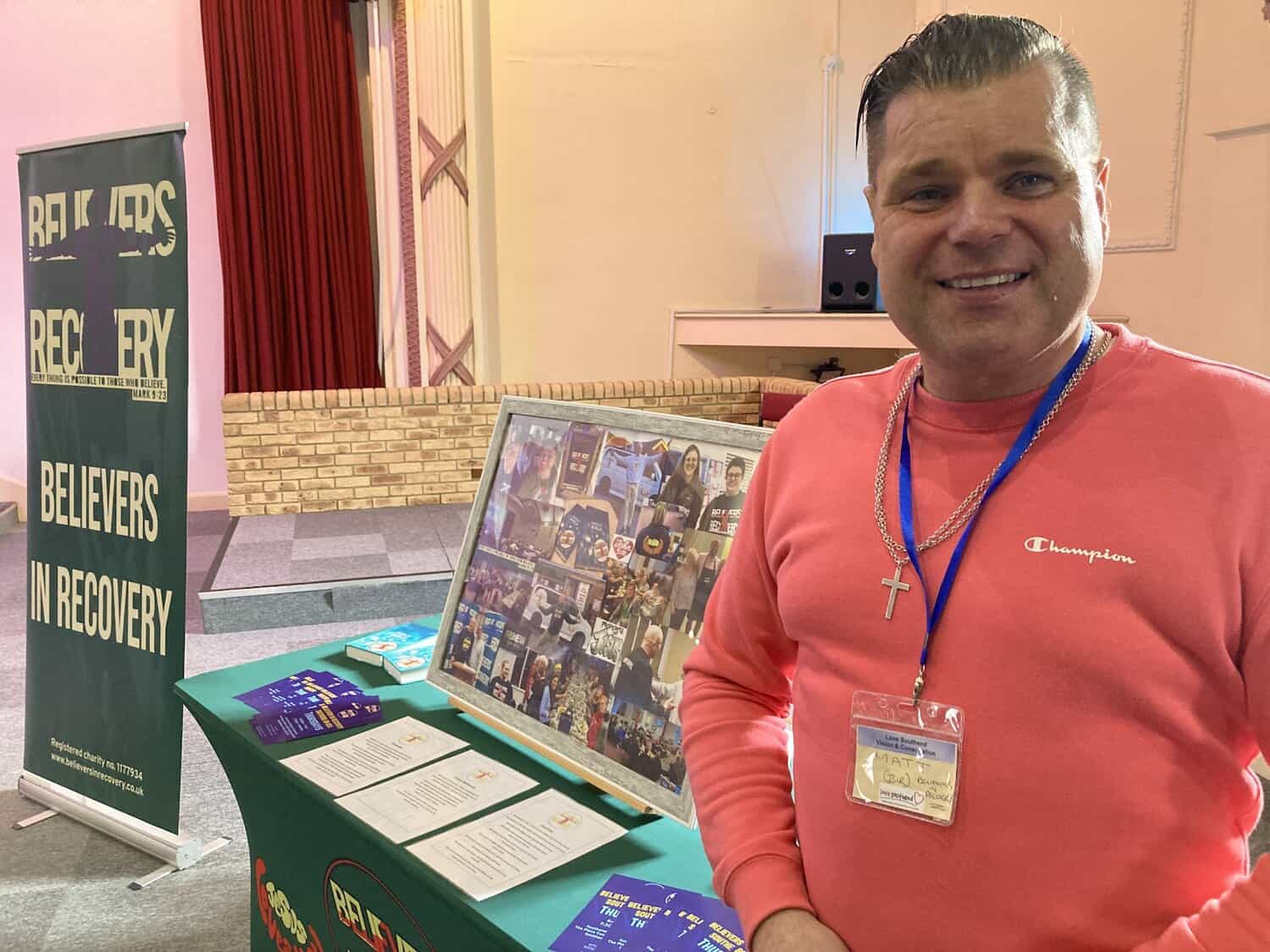 A man stands next to the Believers in Recovery stand an event.